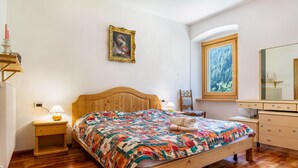 A bedroom with a wooden bed, a nightstand with a lamp, a framed picture on the wall, a window with a view, and a dresser.
