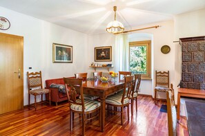 A dining area with a wooden table and chairs, a chandelier, and a window with a view of greenery.