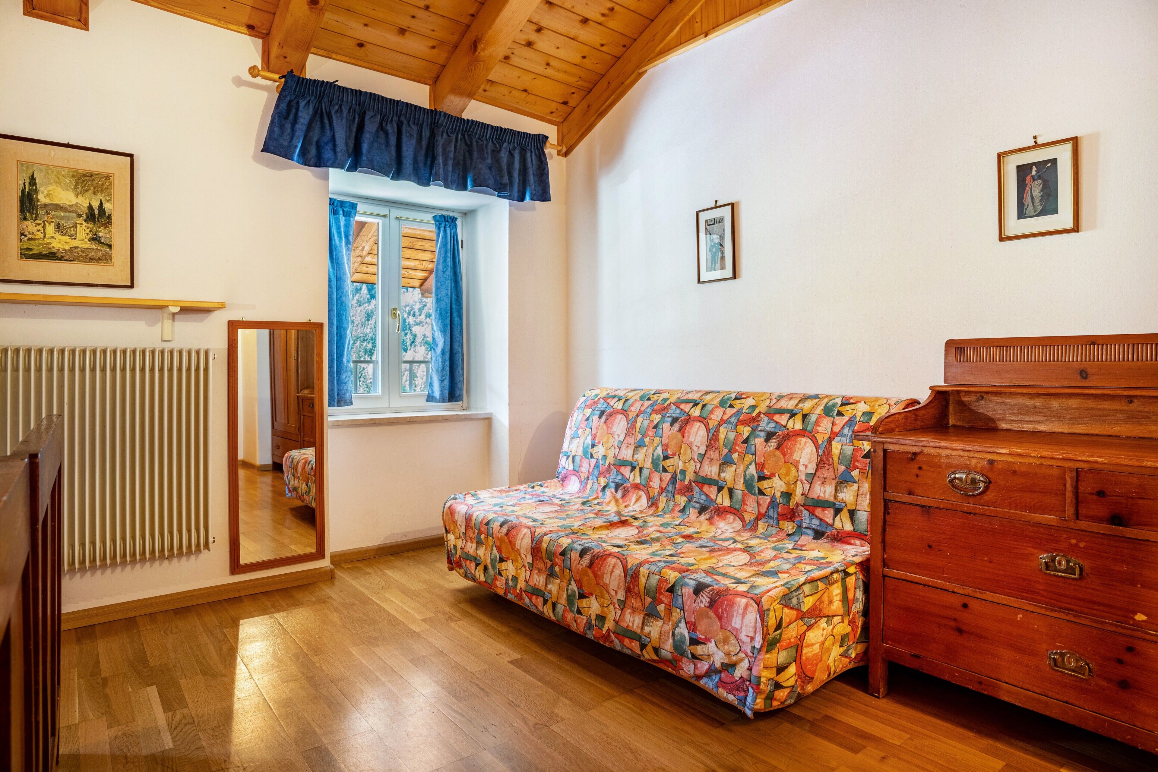 A room with a wooden floor, a colorful sofa, a wooden dresser, a window with blue curtains, and a radiator.