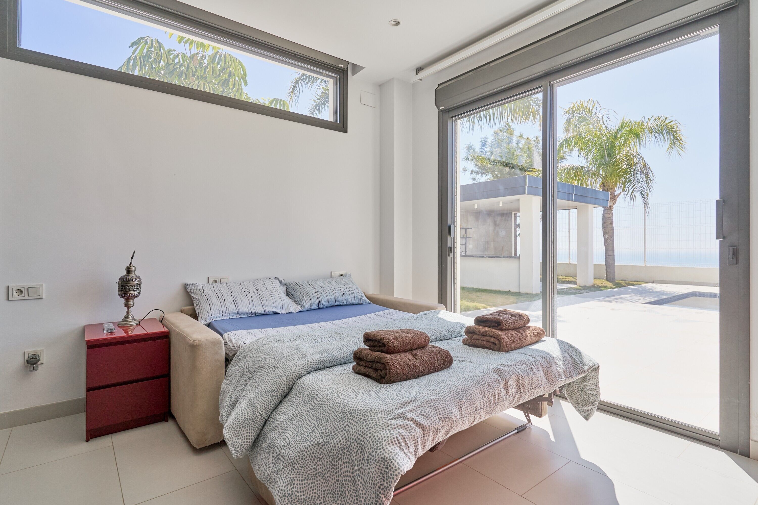 A bedroom with a bed, a nightstand, a lamp, and a view of the outside.