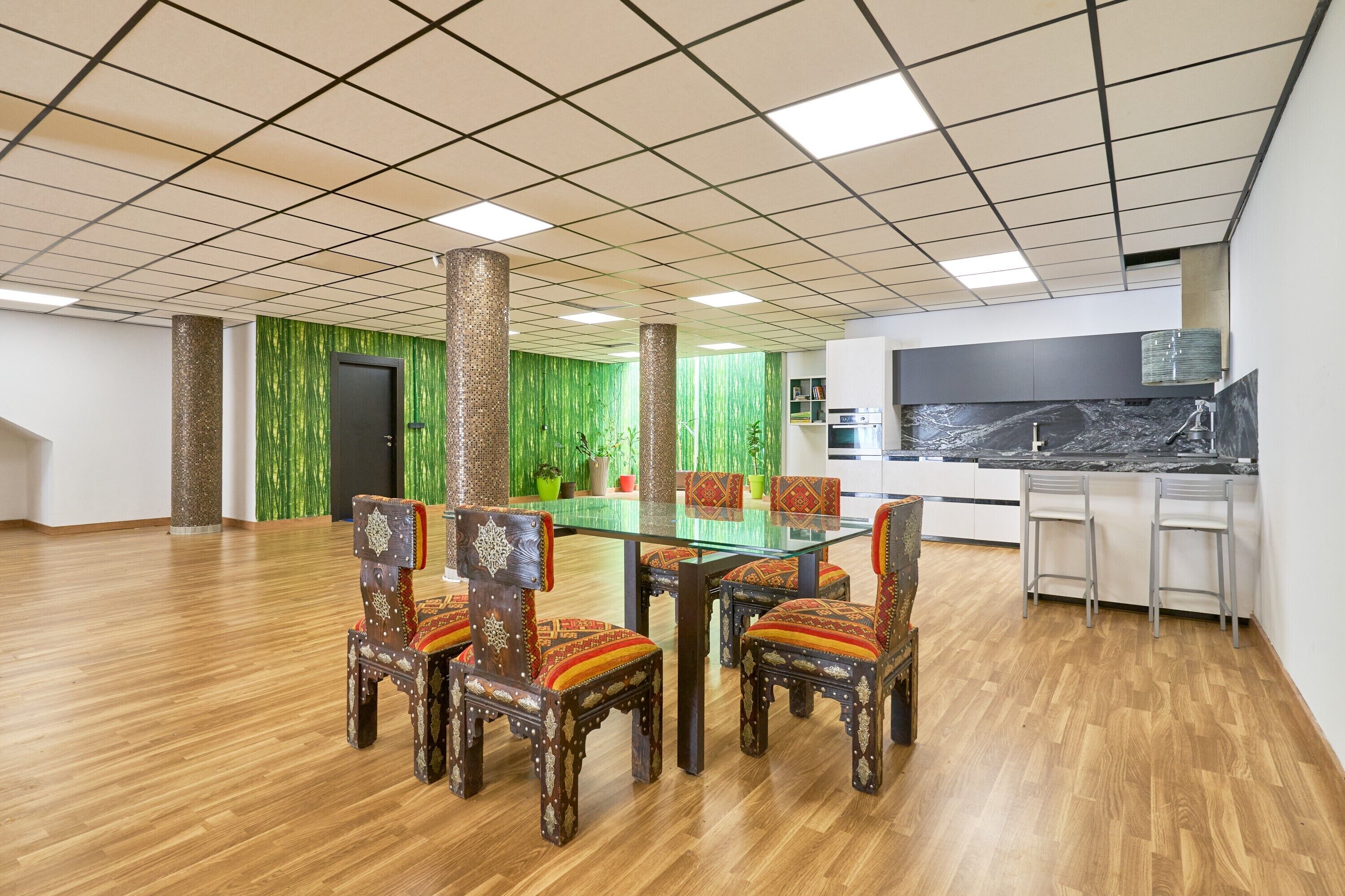 A modern meeting room with a glass table, patterned chairs, a green accent wall, and a kitchen area with marble countertops.