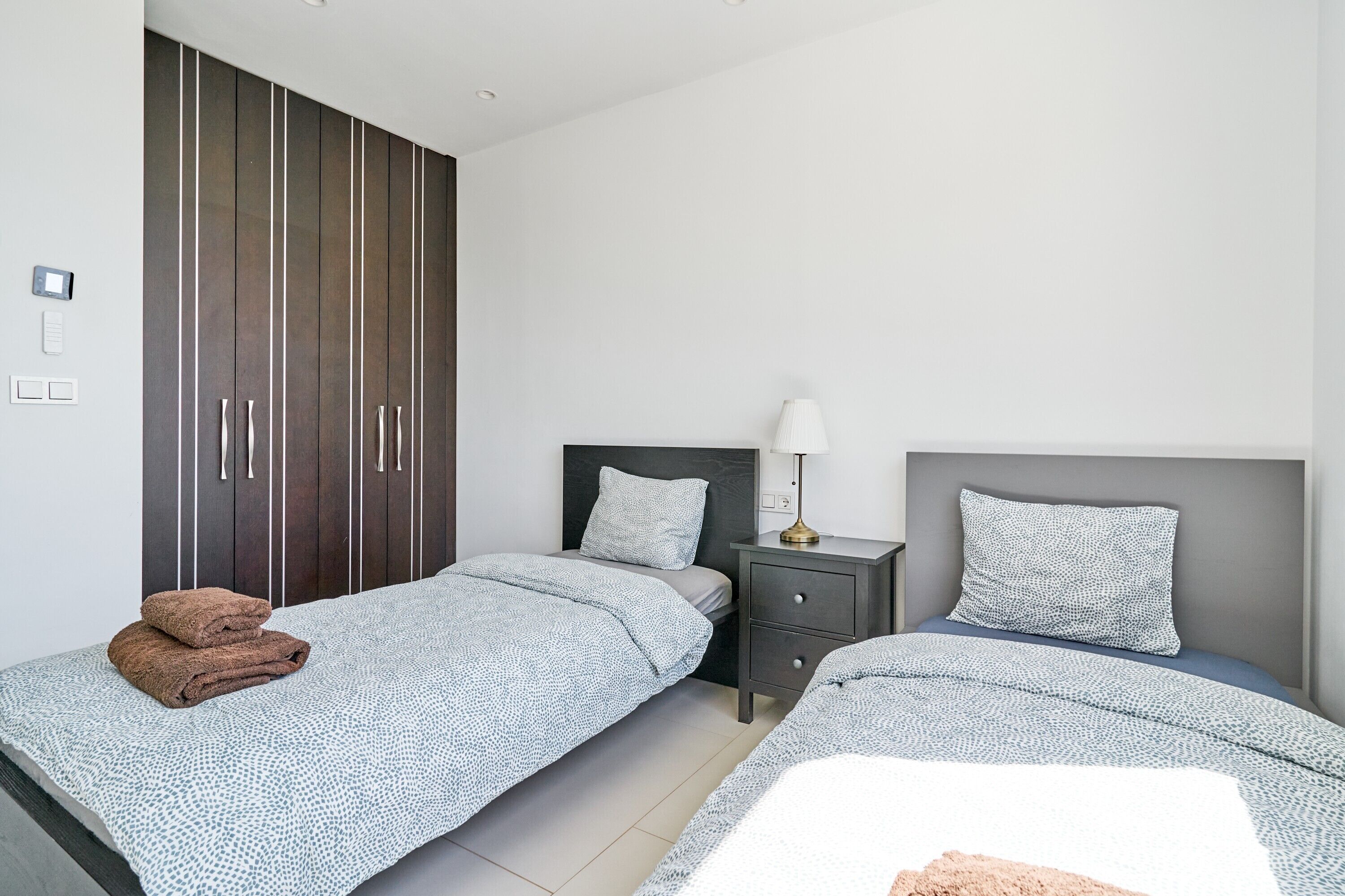 A bedroom with two beds, a wardrobe, a bedside table with a lamp, and a small drawer unit.