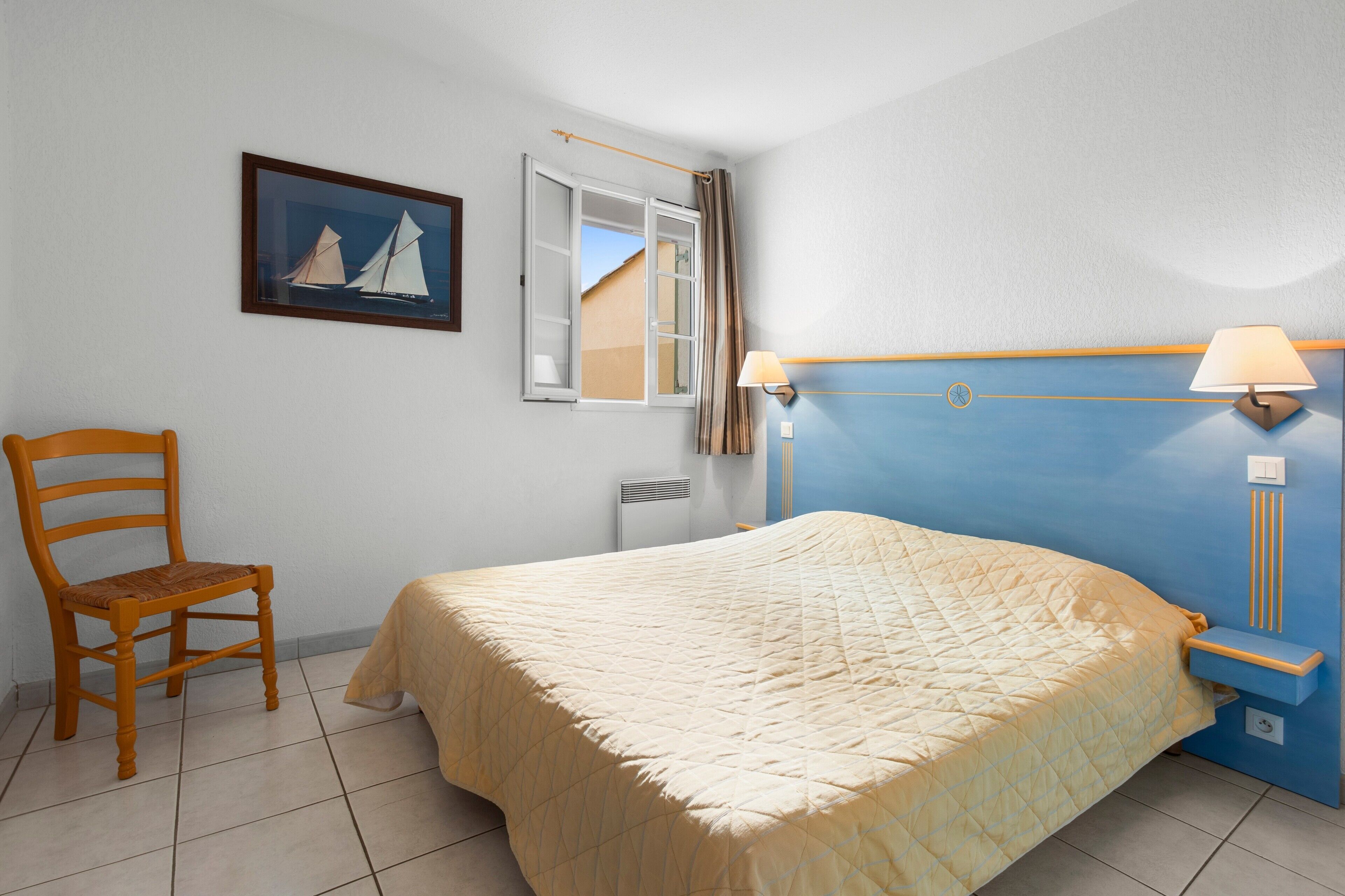 Une chambre à coucher avec un lit, une chaise, une fenêtre avec des rideaux, une lampe fixée au mur et un tableau.