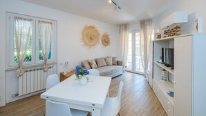 A modern living room with a white sofa, a dining table with chairs, a TV on a shelf, and a large window with curtains. - "Isabel Beach and Bike" Apartment with Private Garden and Wi-Fi (Finale Ligure)