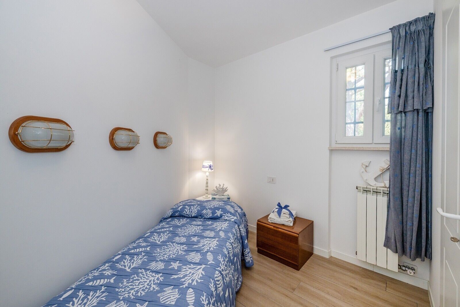 A bedroom with a bed, a nightstand, a window with curtains, and wall-mounted light fixtures.