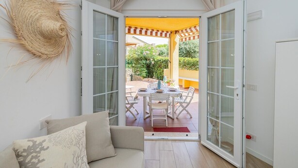 An open door leading to an outdoor patio with a table and chairs, a woven wall decoration, and a white sofa with cushions inside. - "Isabel Beach and Bike" Apartment with Private Garden and Wi-Fi (Finale Ligure)