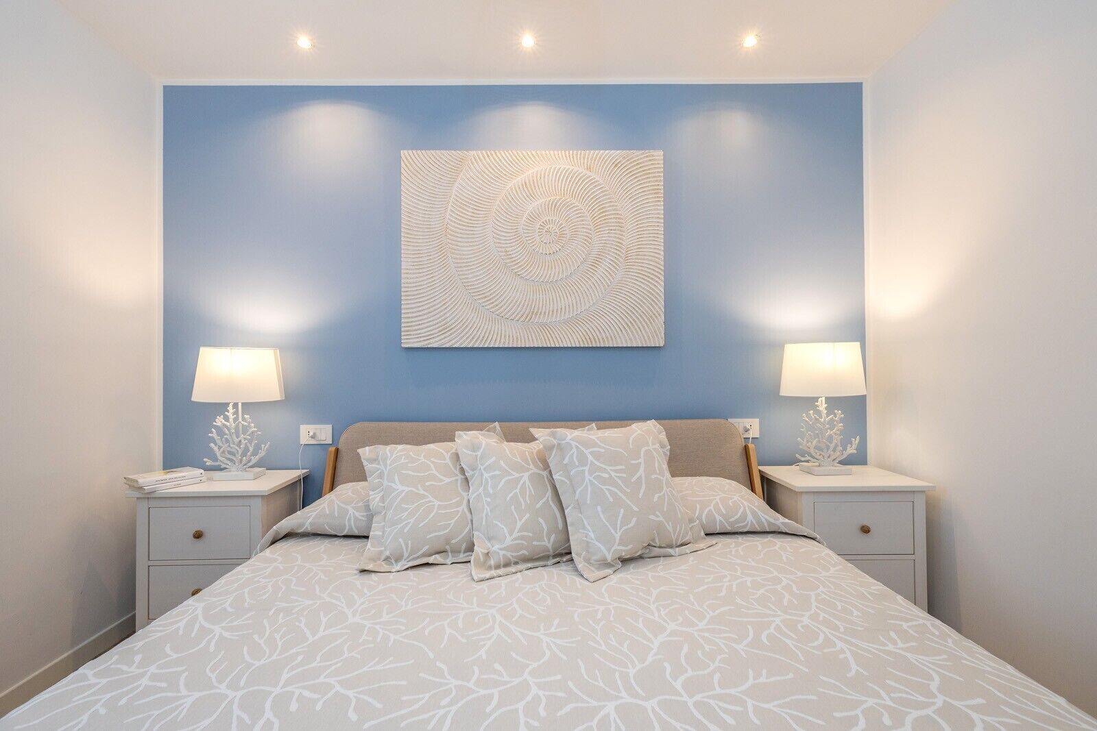 A bedroom with a bed, two bedside tables, a wall-mounted artwork, and two wall-mounted lamps.