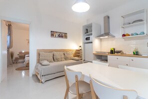 A modern kitchen with white cabinetry, a dining area with a white table and wooden chairs, and a cozy living area with a sofa and patterned cushions.