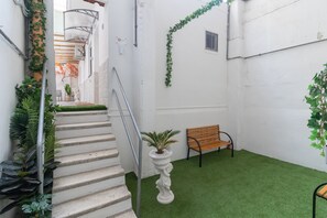 A narrow staircase with a metal handrail, a white wall, a bench, and green artificial grass.