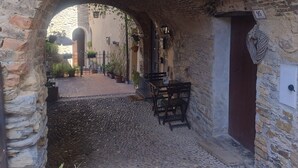 An outdoor stone archway leading to a cobblestone path with a table and chairs, surrounded by potted plants.