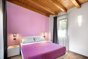 A bedroom with a wooden ceiling, a pink wall, a bed with a purple quilt, bedside tables with lamps, and a window with curtains. - Apartment "Agriturismo Il Borgo - Ulivo" with Private Terrace and Wi-Fi (Villanova d'Albenga)