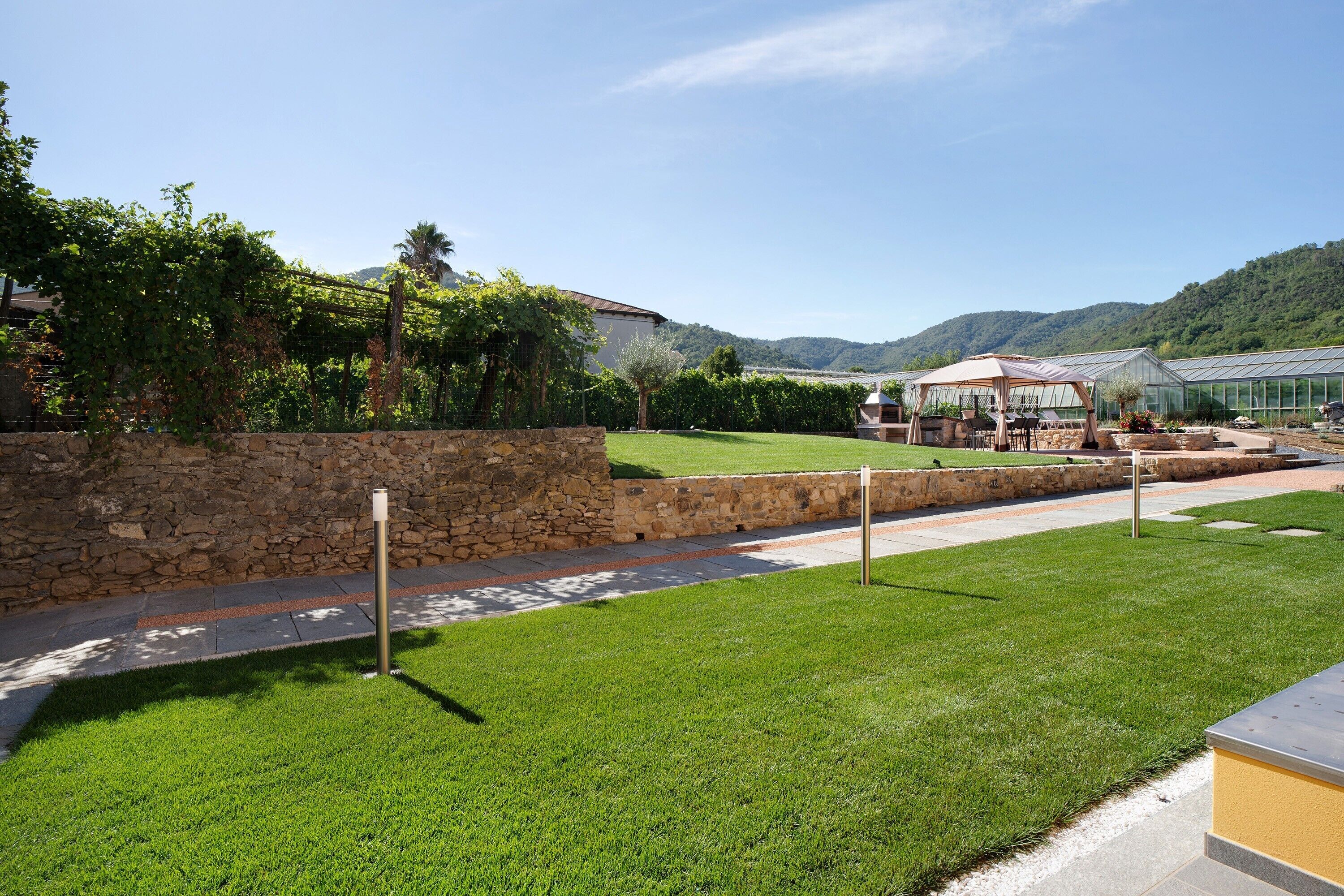 A well-maintained garden with a stone wall, a grassy lawn, and a greenhouse structure.