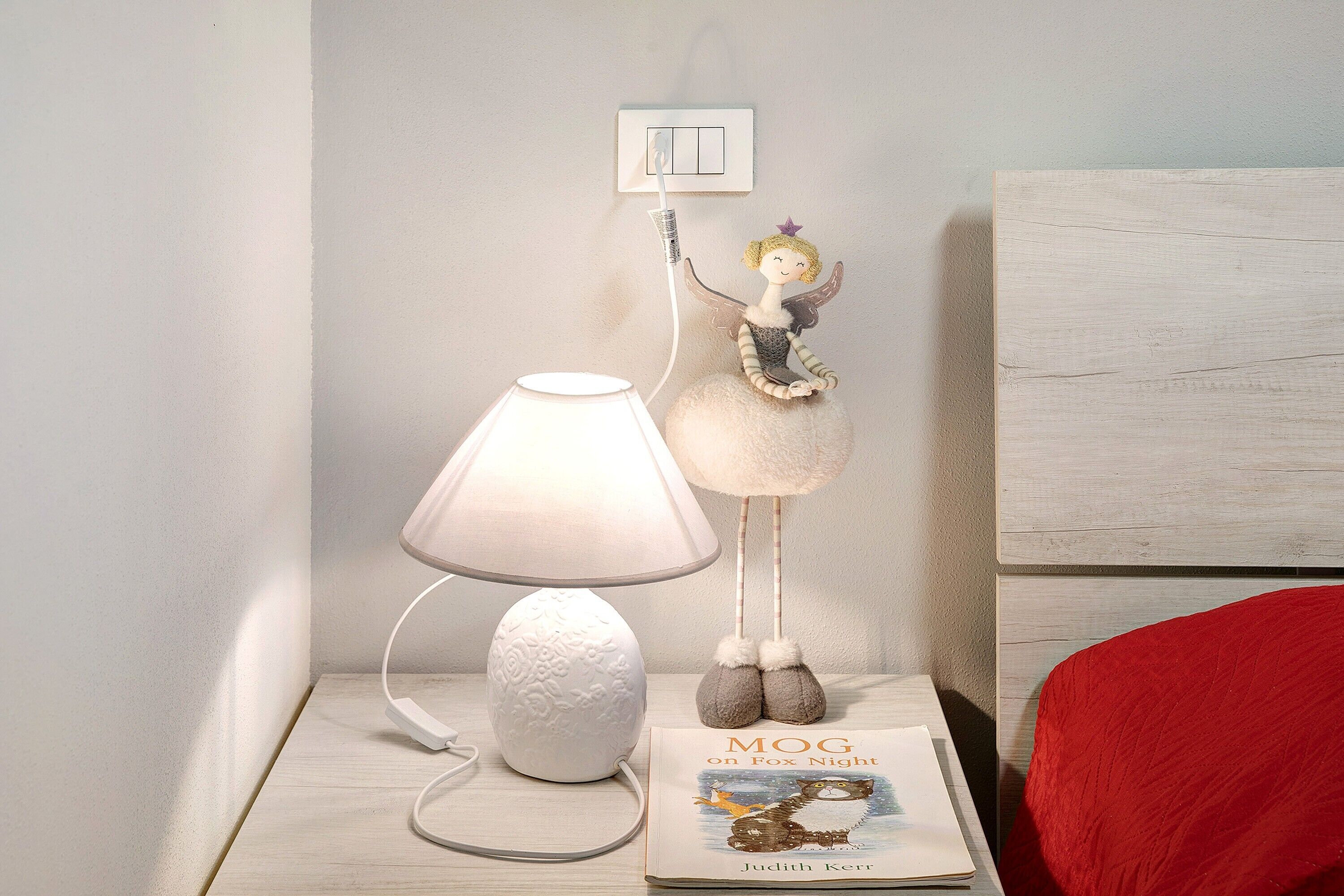 A bedside table with a lamp, a book titled 'MOG on Fox Night' by Judith Kerr, and a fairy figurine.