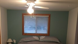 A bedroom with a bed, a ceiling fan, a window with blinds, a nightstand with a lamp, and a wall with a circular pattern.