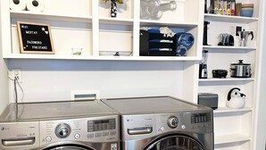 A laundry room with two stainless steel front-loading washers and dryers, a white shelving unit above with various items, and a sign indicating WiFi and password.