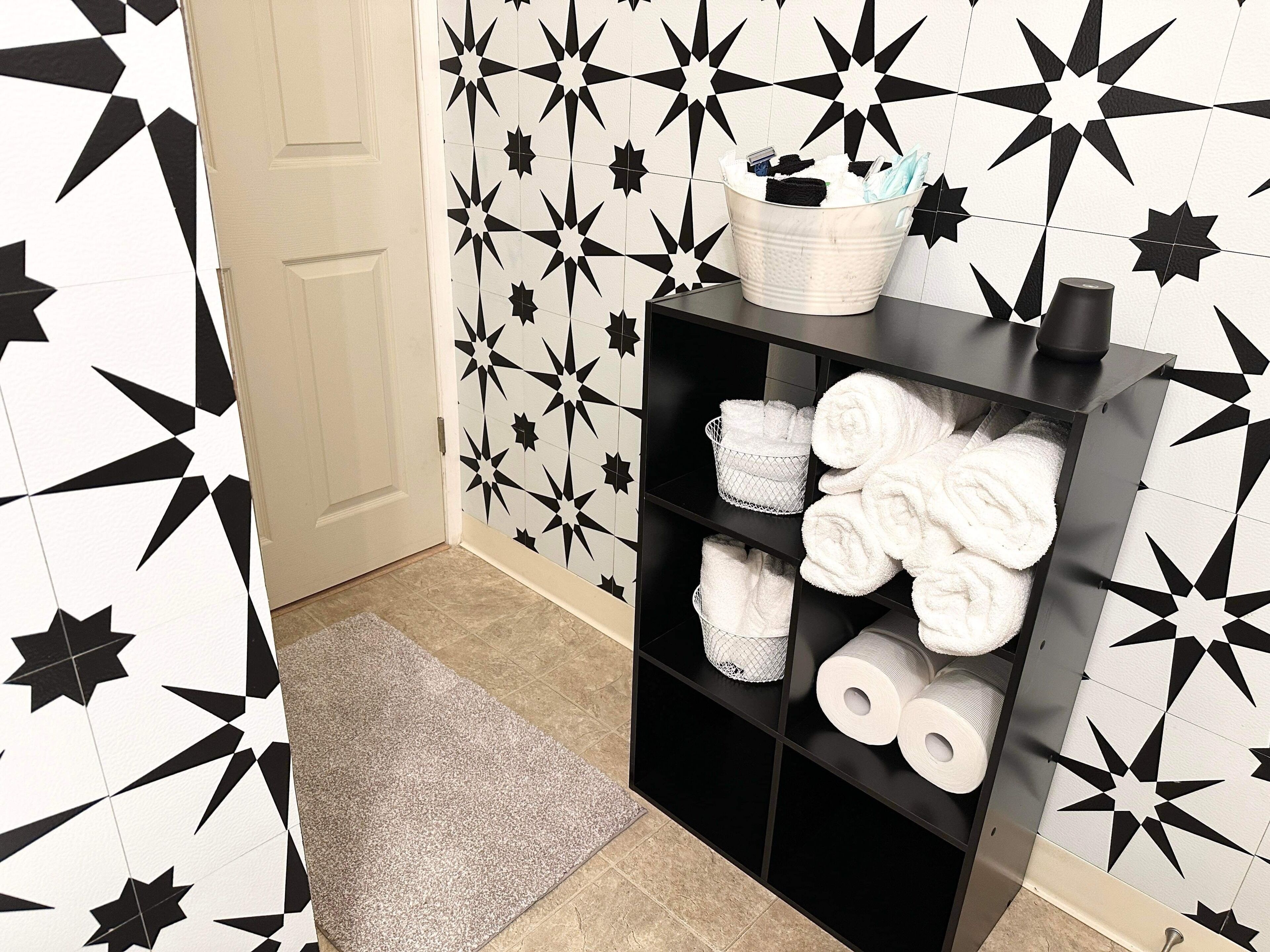 Un baño con una pared con diseño de estrellas en blanco y negro, un estante negro con toallas y papel higiénico, y una alfombra gris en el suelo.