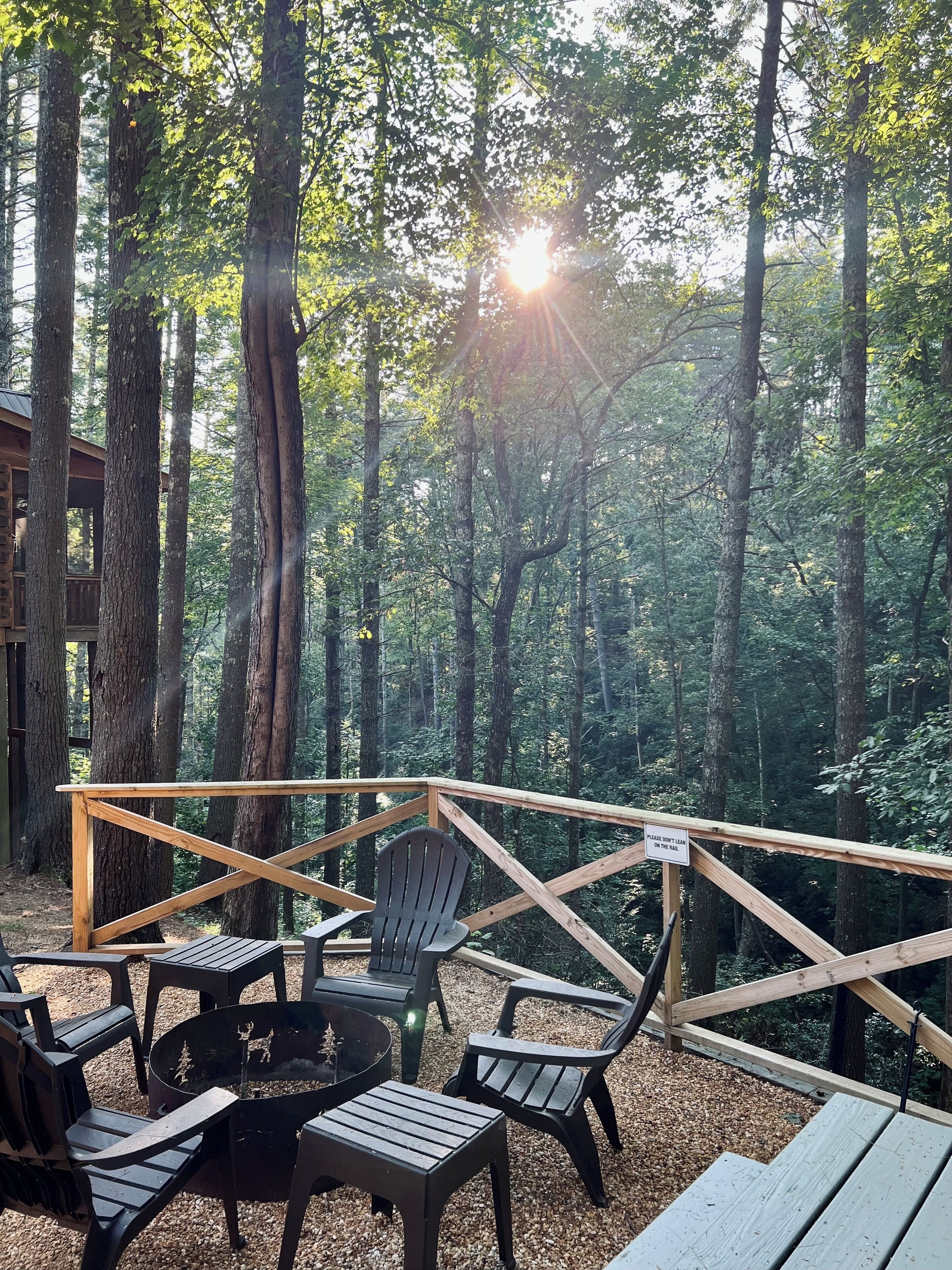 Una terraza de madera con sillas Adirondack y un pozo de fuego, rodeada por un bosque. Hay un cartel que dice 'No apoyarse en la barandilla'.