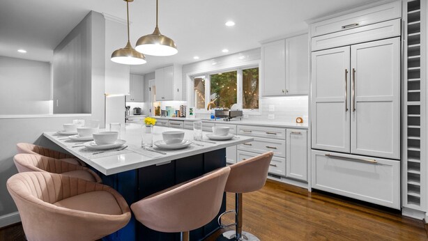 A modern kitchen with white cabinetry, a central island with bar stools, pendant lighting, and a view of the outdoors through large windows. - 6 Bed High End Home w/ EPIC Backyard! (Arlington)