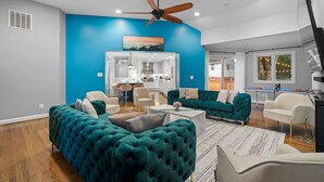 A modern living room with a teal sofa, white armchairs, and a coffee table. There is a ceiling fan, recessed lighting, and a view into the kitchen area. - 6 Bed High End Home w/ EPIC Backyard! (Arlington)