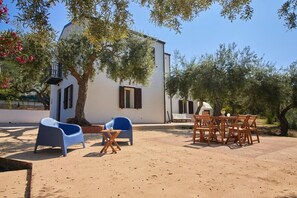A white building with a balcony, surrounded by olive trees and a sandy ground.