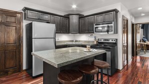 A modern kitchen with dark wood cabinets, a stainless steel refrigerator, a microwave, and an oven. There is a granite countertop with a coffee maker and a small island with stools. - Century Cafe Flat (Kewaskum)