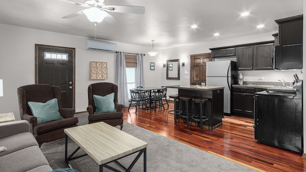 A modern kitchen with dark cabinets, a central island, and a bar area. There is a dining area with a table and chairs, and a living room with a sofa and armchairs. - Century Cafe Flat (Kewaskum)