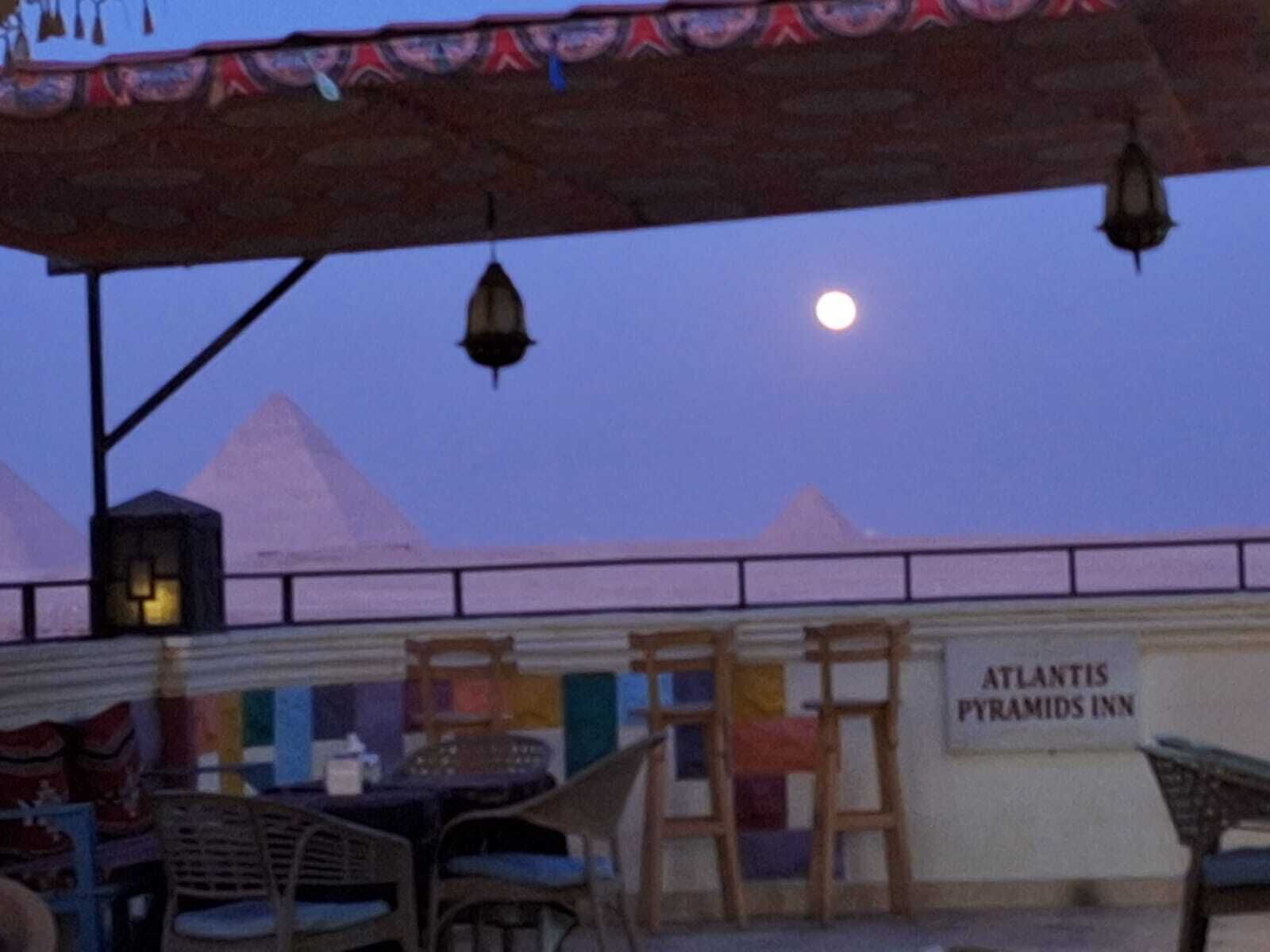 Vista del Atlantis Pyramids Inn con un letrero, área de asientos al aire libre y un cielo nocturno con la luna.