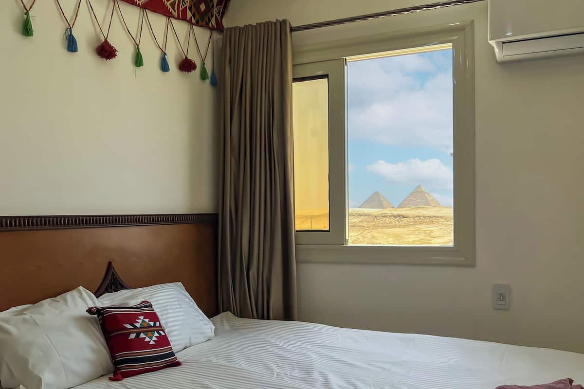 Ein Hotelzimmer mit einem Bett, einem Fenster mit Blick auf Pyramiden und einer Wand mit hängenden Quasten.
