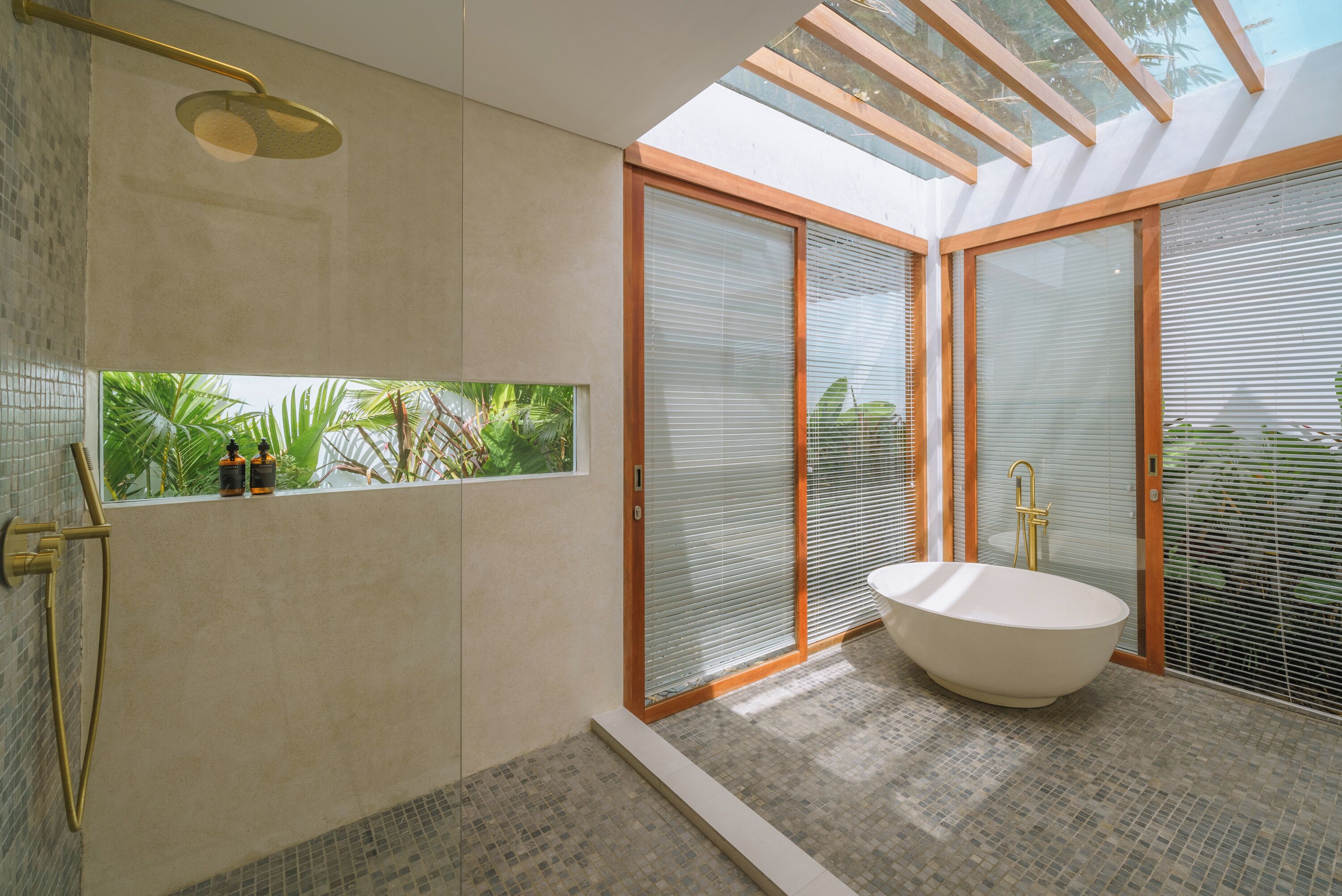 A modern bathroom with a freestanding bathtub, a shower with a gold fixture, and a view of greenery through a window.