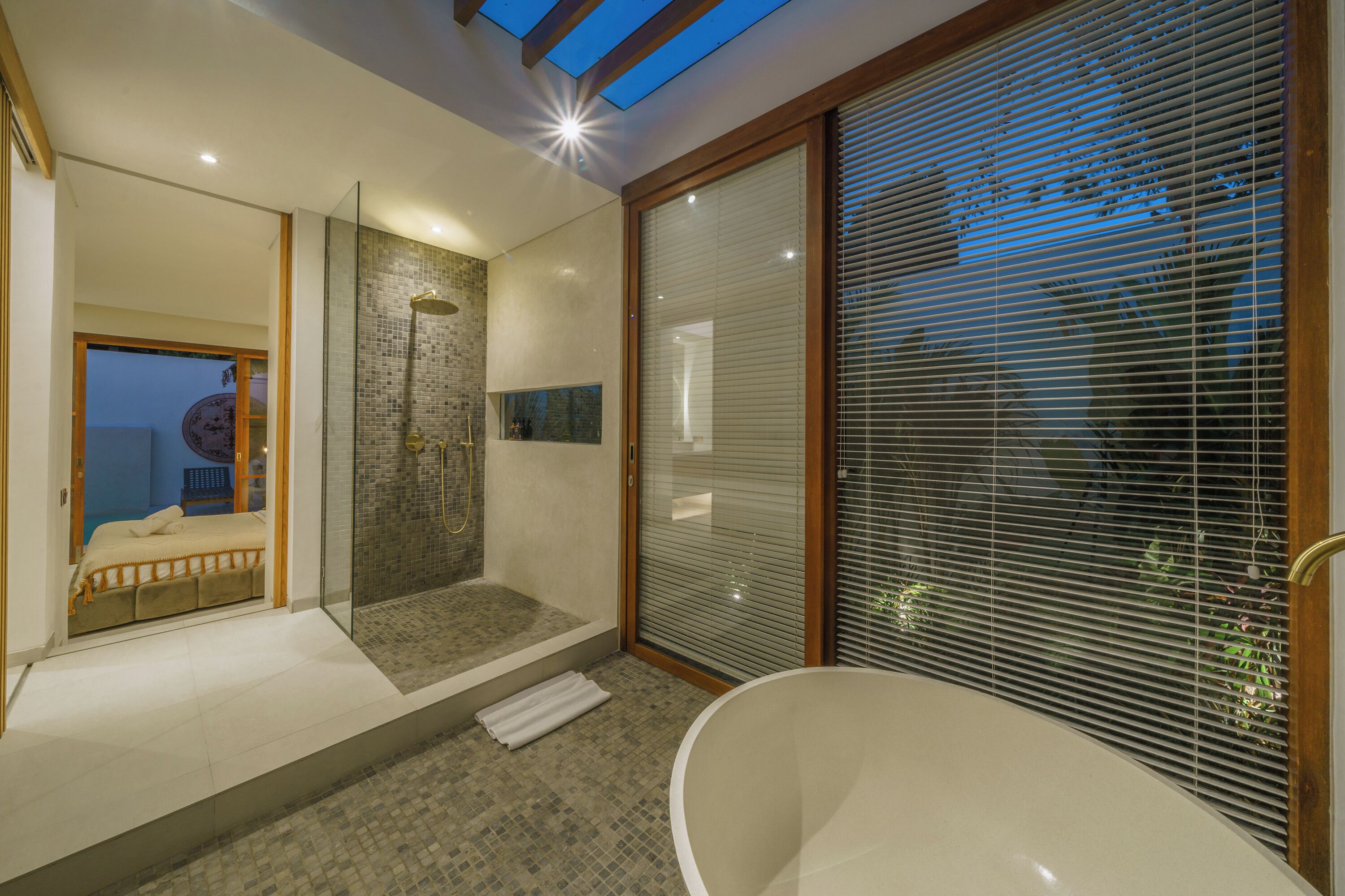 A modern bathroom with a glass-enclosed shower, a freestanding bathtub, and a large window with blinds.