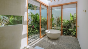 A modern bathroom with a freestanding bathtub, large sliding glass doors, and lush tropical plants. - Villa Cocoon - Gaia (Canggu)