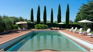 A rectangular pool with clear water, surrounded by white lounge chairs and umbrellas, with tall cypress trees in the background.