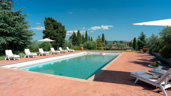 A rectangular pool with white lounge chairs and umbrellas, surrounded by greenery and a clear sky.