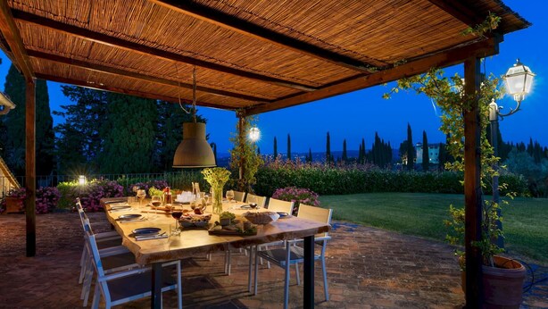 An outdoor dining area with a wooden table set for a meal, surrounded by chairs, under a thatched roof.
