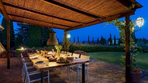An outdoor dining area with a wooden table set for a meal, surrounded by chairs, under a thatched roof. - Villa I Levrieri 8 Emma Villas (Barberino Tavarnelle)