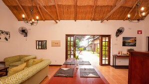 A spacious room with a wooden ceiling, a dining table set with placemats and cutlery, a sofa with patterned cushions, and a view through an open door to an outdoor area. - 2 bed Apartment With Pool - 2126 (Victoria Falls)