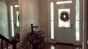 A hallway with a wooden floor, a rug, a chandelier, a door with a wreath, and a mirror reflecting the hallway. - The Indigo House- Located in the heart of Historic Hannibal, Missouri  (Hannibal)