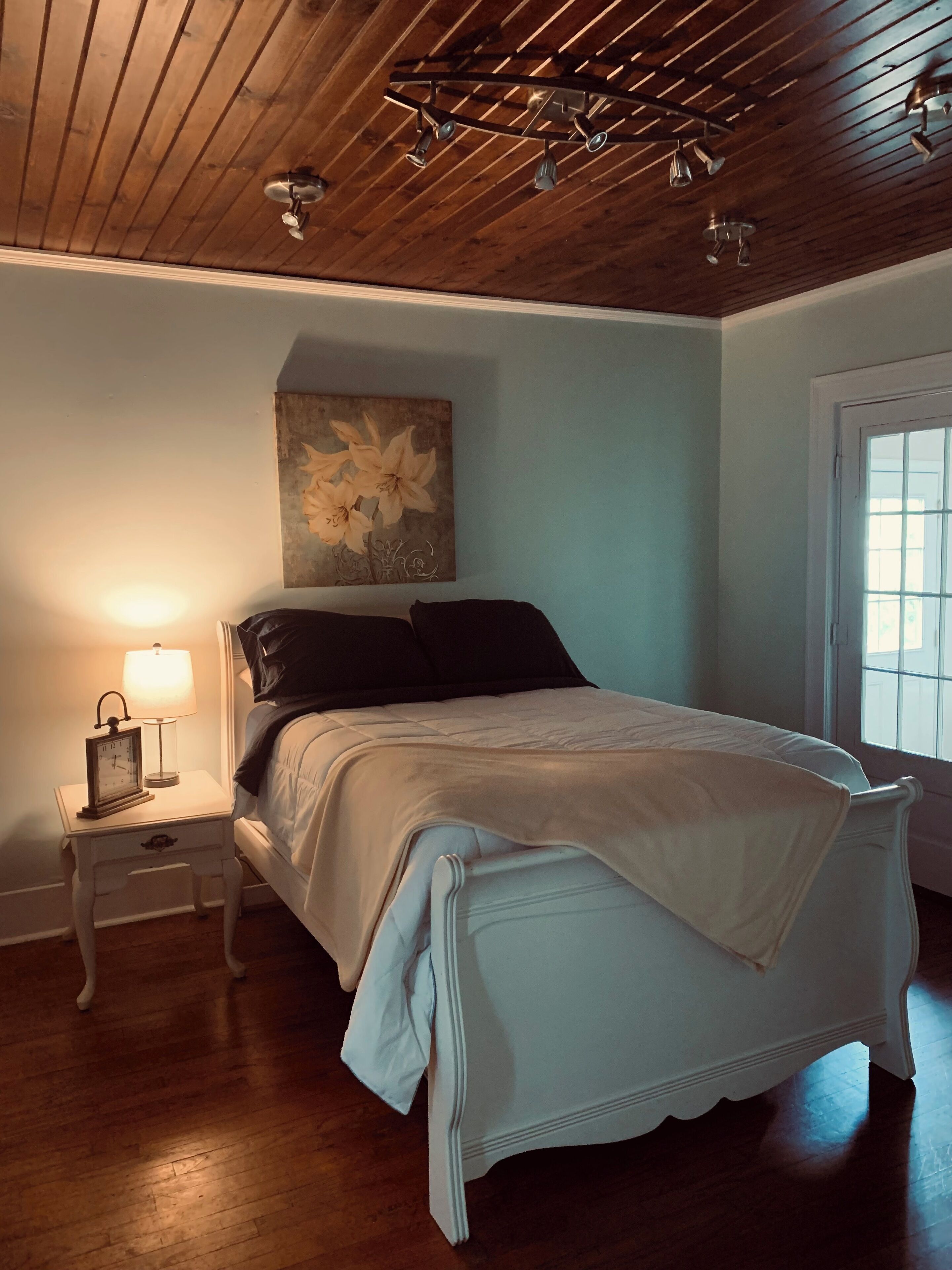 A bedroom with a wooden ceiling, a bed with white bedding, a nightstand with a lamp, and a floral painting on the wall.