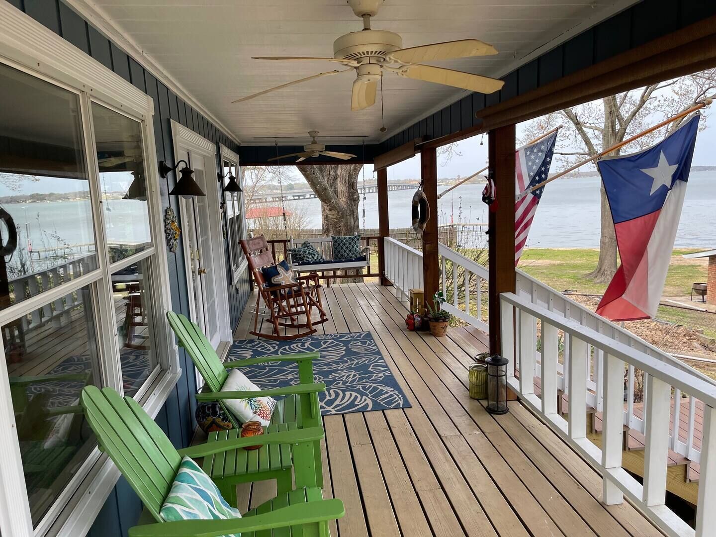 A covered porch with a view of a body of water, a wooden deck, rocking chairs, and a flag.