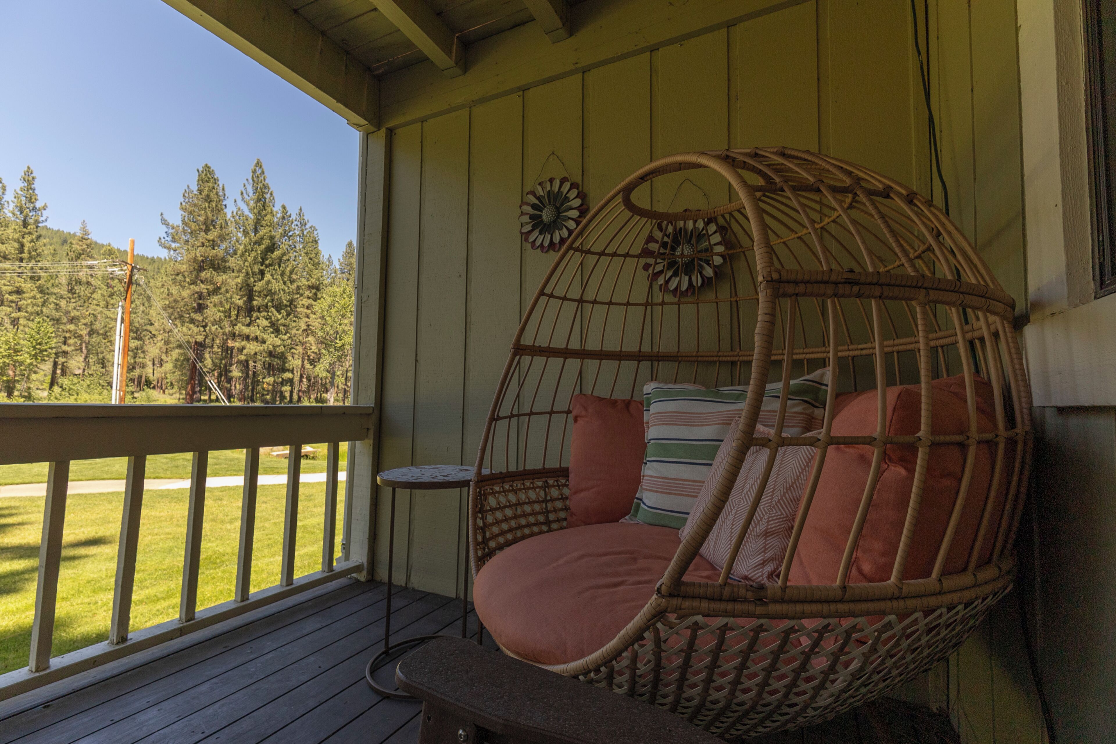 Una silla de mimbre con cojines sobre una terraza de madera, una mesita y vistas a un paisaje verde.