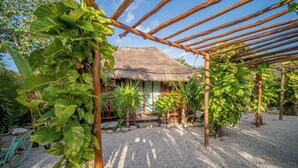 Une construction à toit de chaume, surmontée d’une pergola en bois, entourée de gravier et de plantes tropicales.