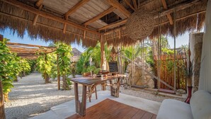 Un espace extérieur aménagé dans un style rustique, avec une table en bois et un banc rembourré, entouré de plantes tropicales et d’un mur en pierre.