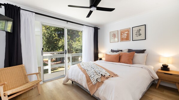 A bedroom with a bed, a chair, a ceiling fan, a nightstand, and a view of the outdoors. - Barney's Beach House - A Haven of Comfort and Style (Narrawallee)
