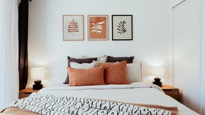 A neatly made bed with a patterned blanket, a headboard with multiple pillows, bedside tables with lamps, and three framed botanical prints on the wall. - Barney's Beach House - A Haven of Comfort and Style (Narrawallee)