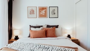 A neatly made bed with a patterned blanket, a headboard with multiple pillows, bedside tables with lamps, and three framed botanical prints on the wall. - Barney's Beach House - A Haven of Comfort and Style (Narrawallee)