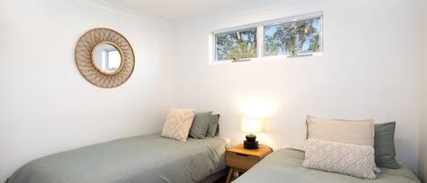 Un dormitorio con dos camas individuales, una mesita de noche con lámpara, un espejo redondo y una ventana con vistas a árboles.