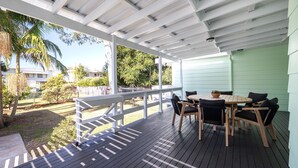 A covered outdoor patio with a wooden table and chairs, a white railing, and a view of a residential area with houses and trees. - Barney's Beach House - A Haven of Comfort and Style (Narrawallee)
