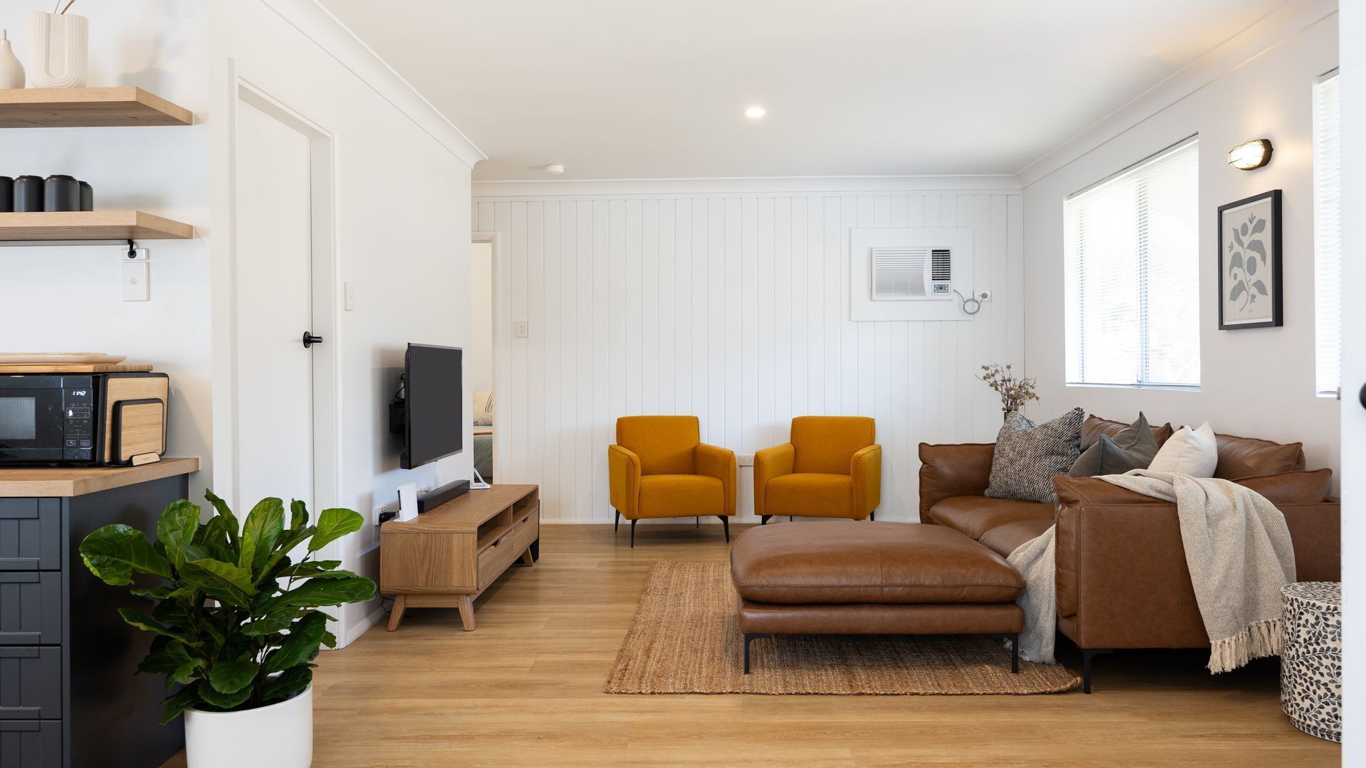 Un salon moderne avec un plancher en bois, un canapé en cuir marron, deux fauteuils de couleur moutarde, un téléviseur posé sur un meuble en bois et une plante en pot.