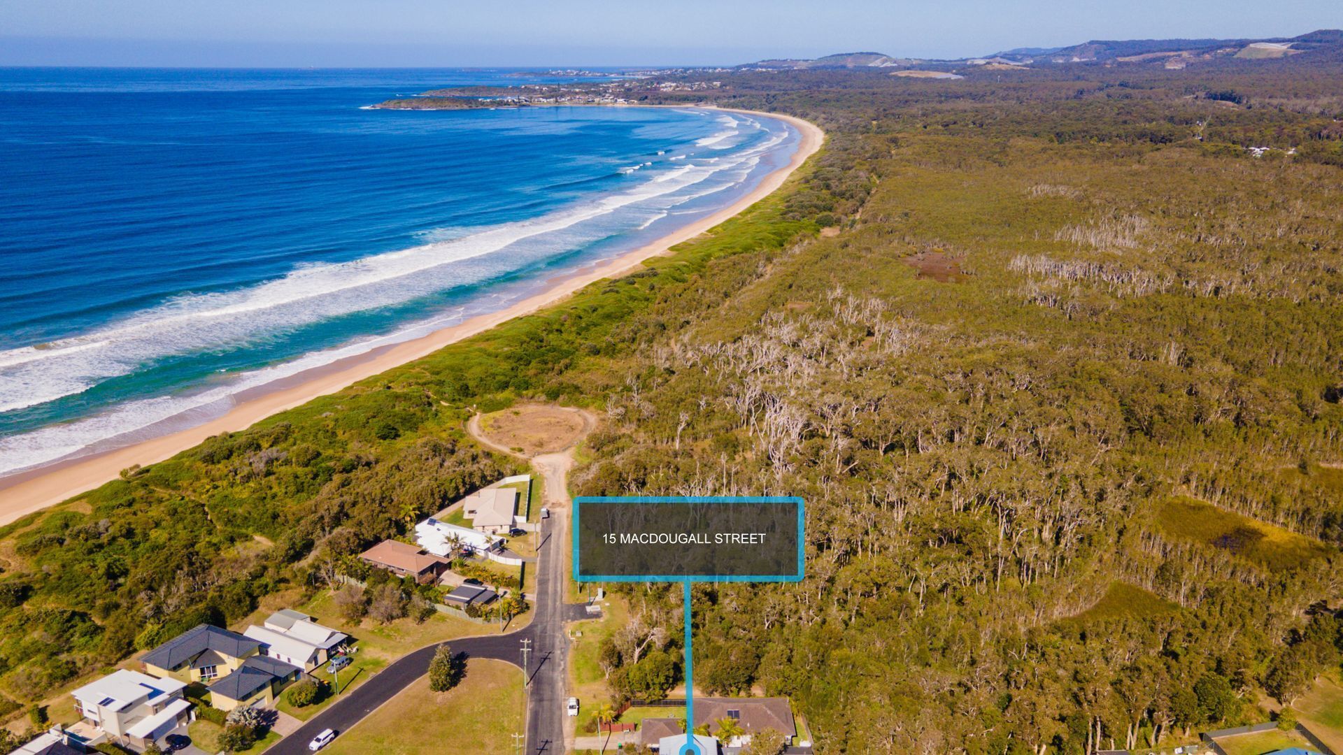 An aerial view of a coastal property with a beach, dense forest, and a road leading to a street named 15 Macdougall Street.