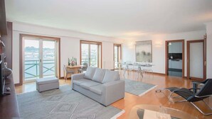 Ein modernes Wohnzimmer mit Sofa, Esstisch und Fernseher. Ein Balkon bietet Blick auf die Stadt.